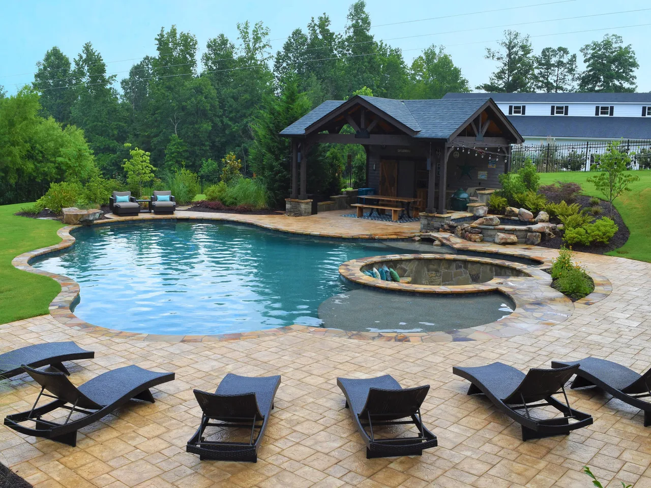 Resort-style backyard with blue water pool, attached spa, paver deck, lounge chairs, and covered pavilion with trees in background.