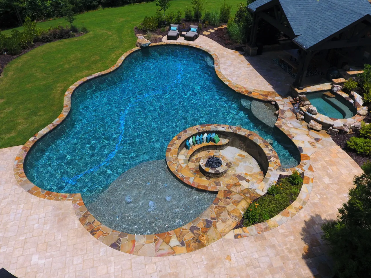 Aerial view of freeform swimming pool with spa and sun shelf surrounded by travertine deck in Alpharetta, Georgia