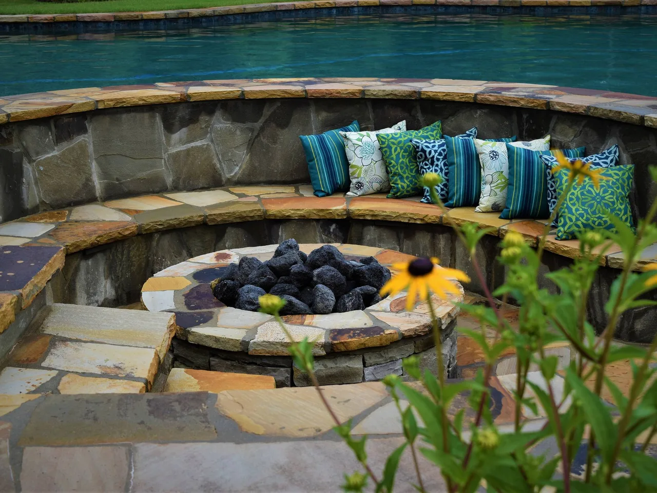 Sunken fire pit with seating ledges and colorful outdoor pillows, stone surround with lit fire bowl, pool visible beyond in Alpharetta, GA