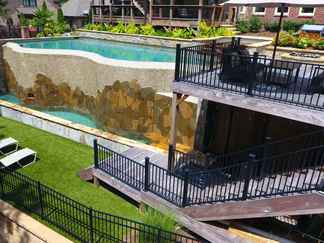 Vanishing edge pool with stacked stone water feature and turquoise water, black metal railings, manicured lawn, and brick home in background