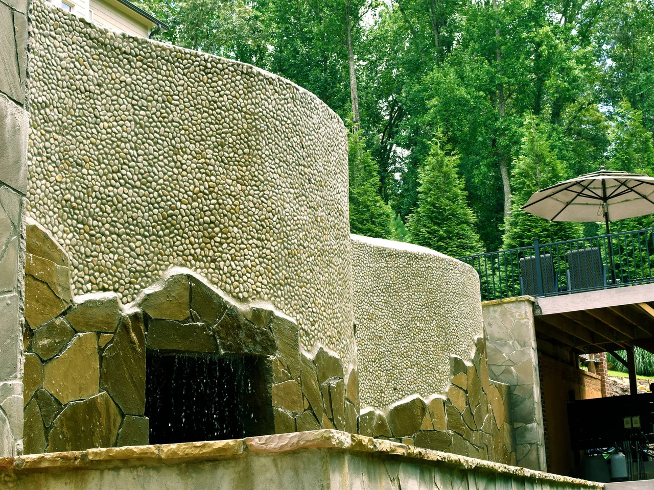 Curved stacked stone water feature with sheer descent into vanishing edge pool, surrounded by tall trees and patio umbrella