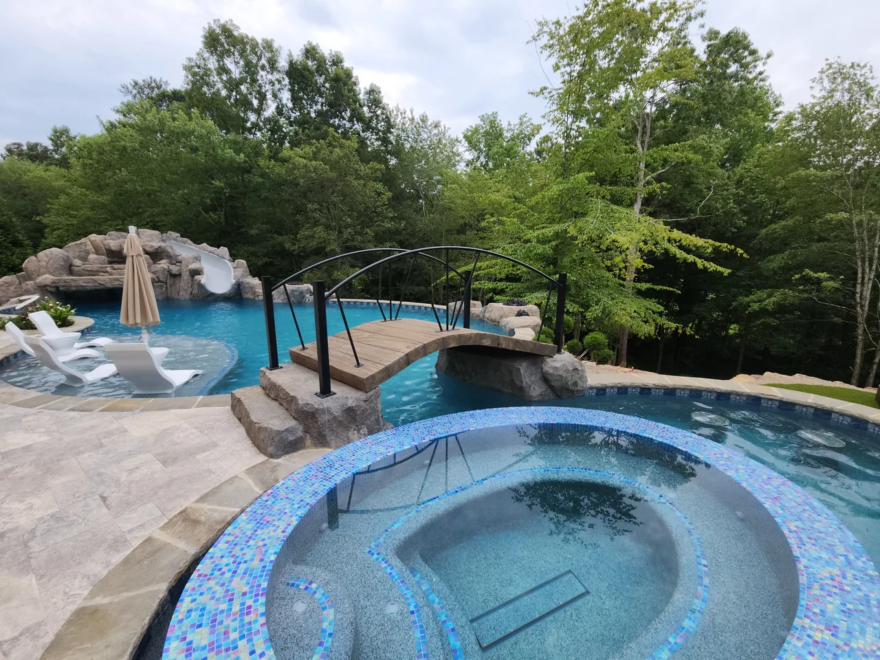 Round spa with blue tile surround, arched bridge, grotto pool, and wooded backdrop in Atlanta