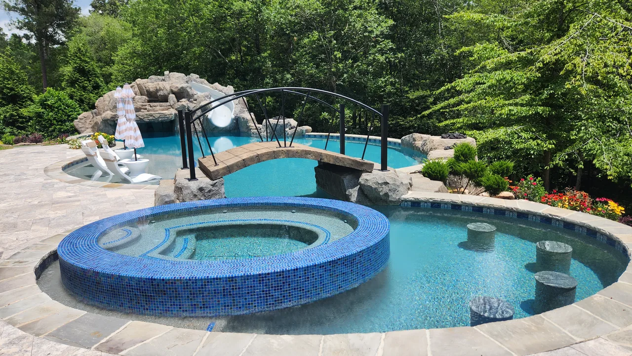 Round spa with blue tile surround, stone bridge, grotto with slide, and multi-level pool with beach entry steps surrounded by mature trees