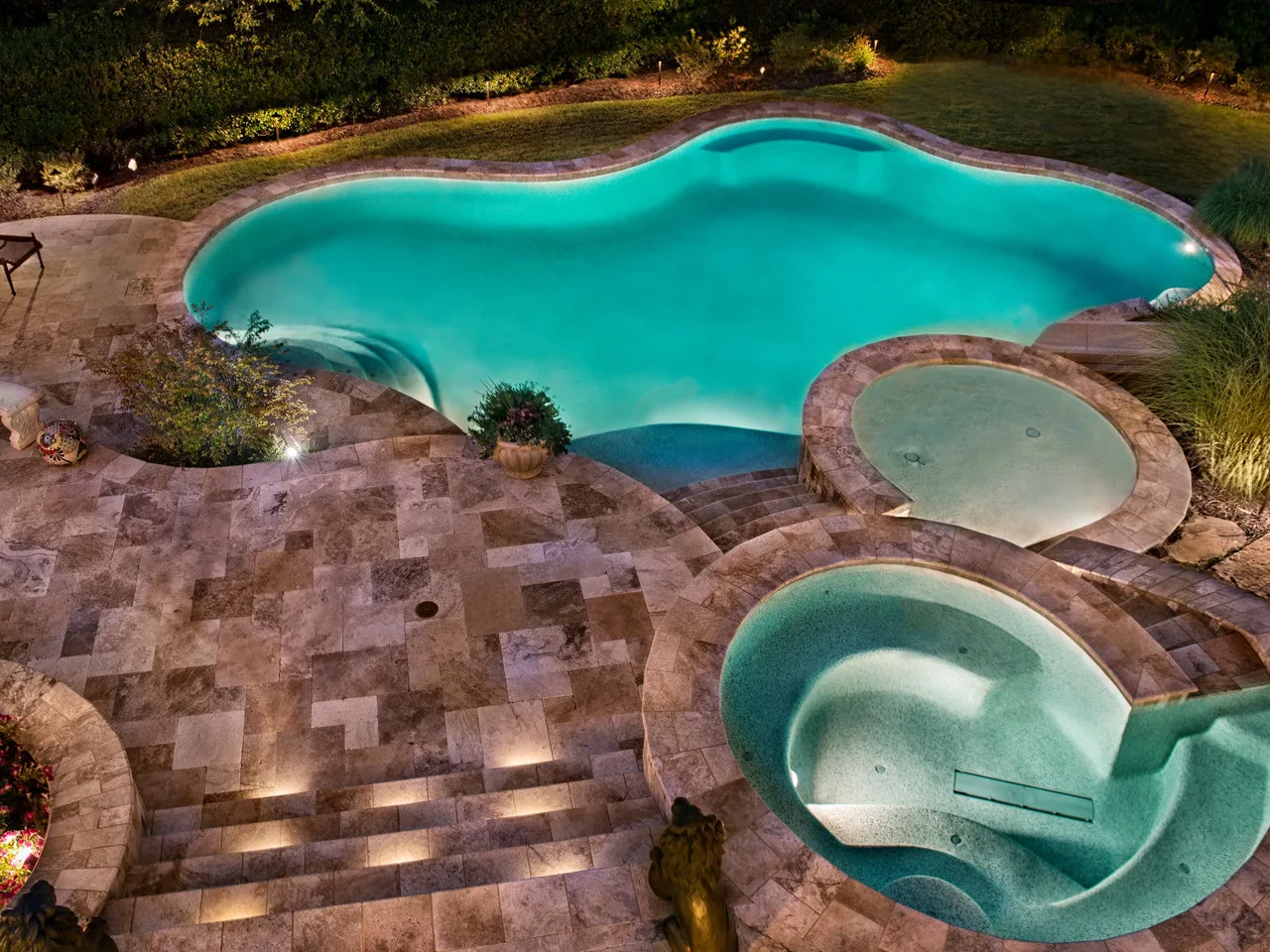Aerial view of freeform pool with turquoise water, travertine deck, two spas, and landscape lighting at dusk in Johns Creek, GA