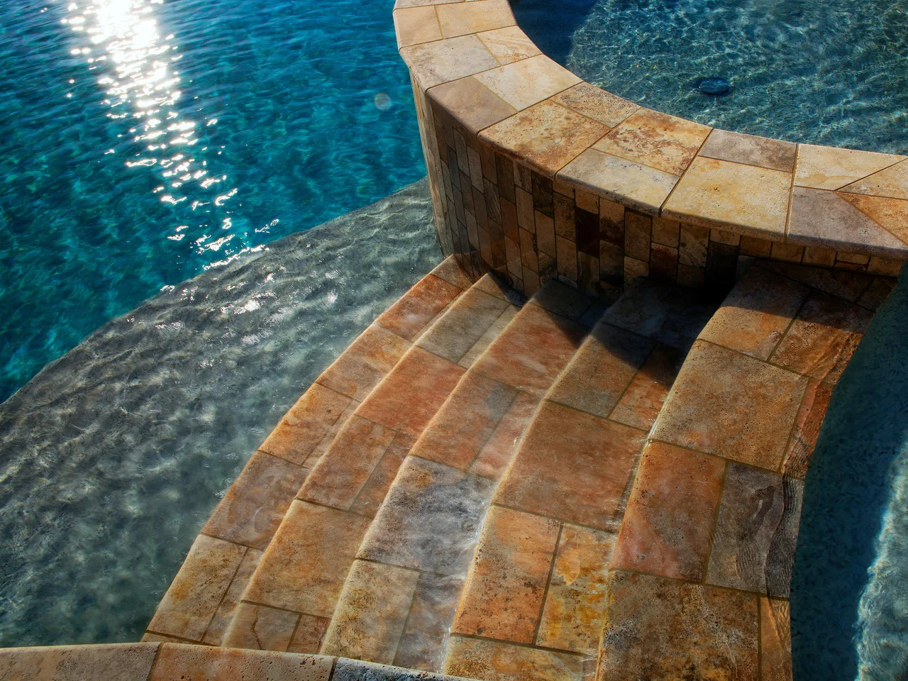 Curved travertine deck and sun shelf beside crystal blue pool water with cascading step waterfall in Johns Creek, GA
