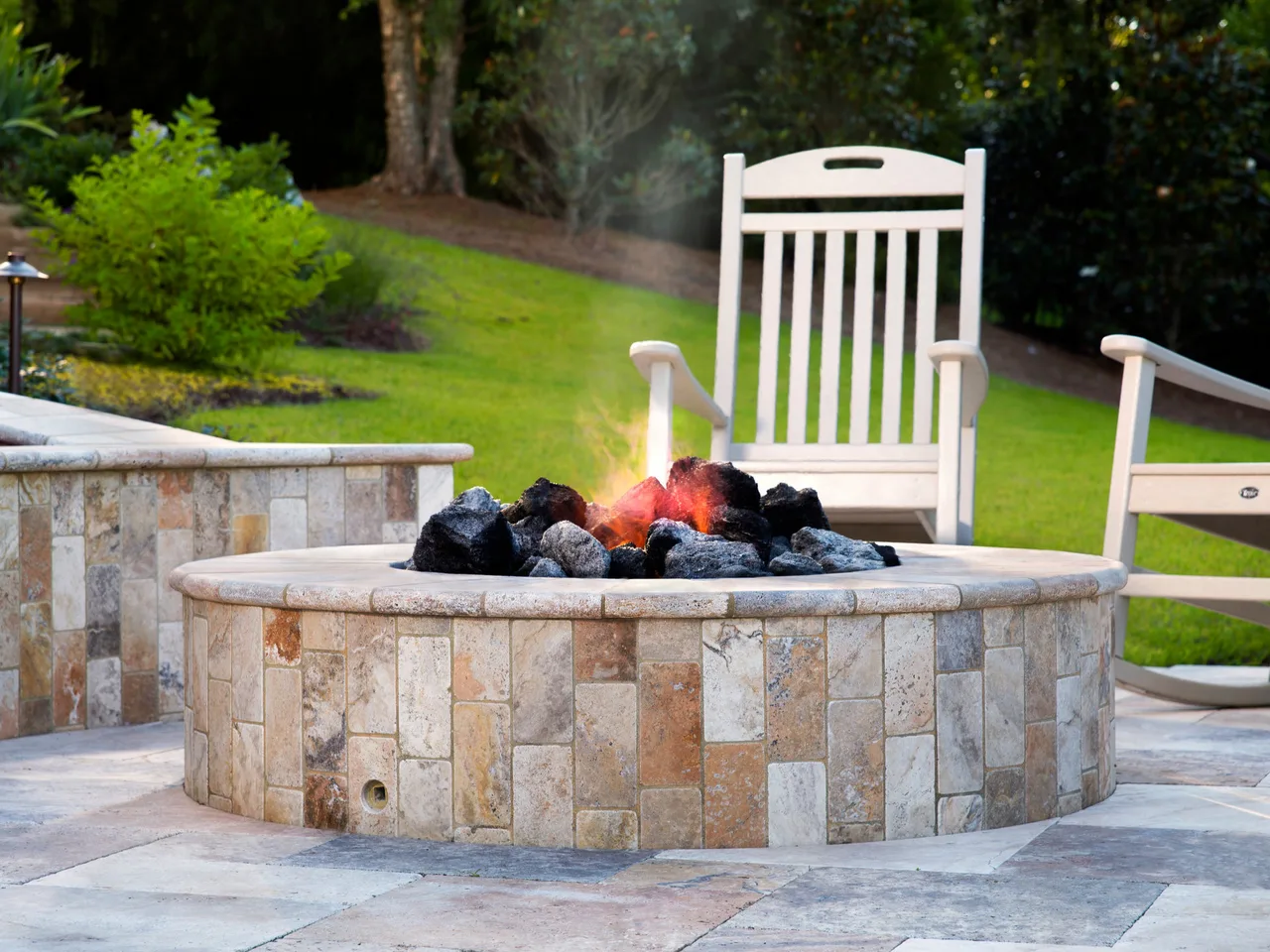 Stone fire bowl with glowing embers and dark glass rocks on travertine patio beside white Adirondack chairs and green lawn in Johns Creek, GA