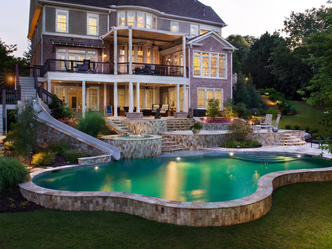 Illuminated freeform pool with travertine coping and spa at dusk, three-tier stone decking with waterslide, and brick home with lit windows in Johns Creek, GA