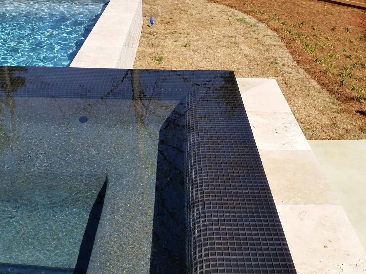 Dark tile scupper edge with white coping bordering crystal blue pool water and sandy deck area
