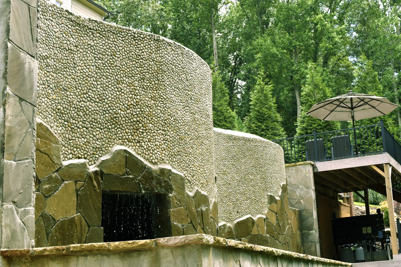 Stone spa with sheer descent waterfall, curved travertine walls, pergola pavilion with umbrella, and mature trees