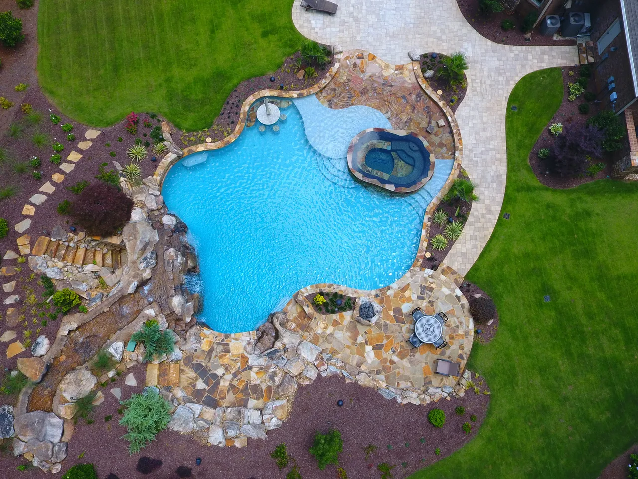 Aerial view of freeform pool with cave grotto, beach entry, and travertine deck surrounded by manicured lawn