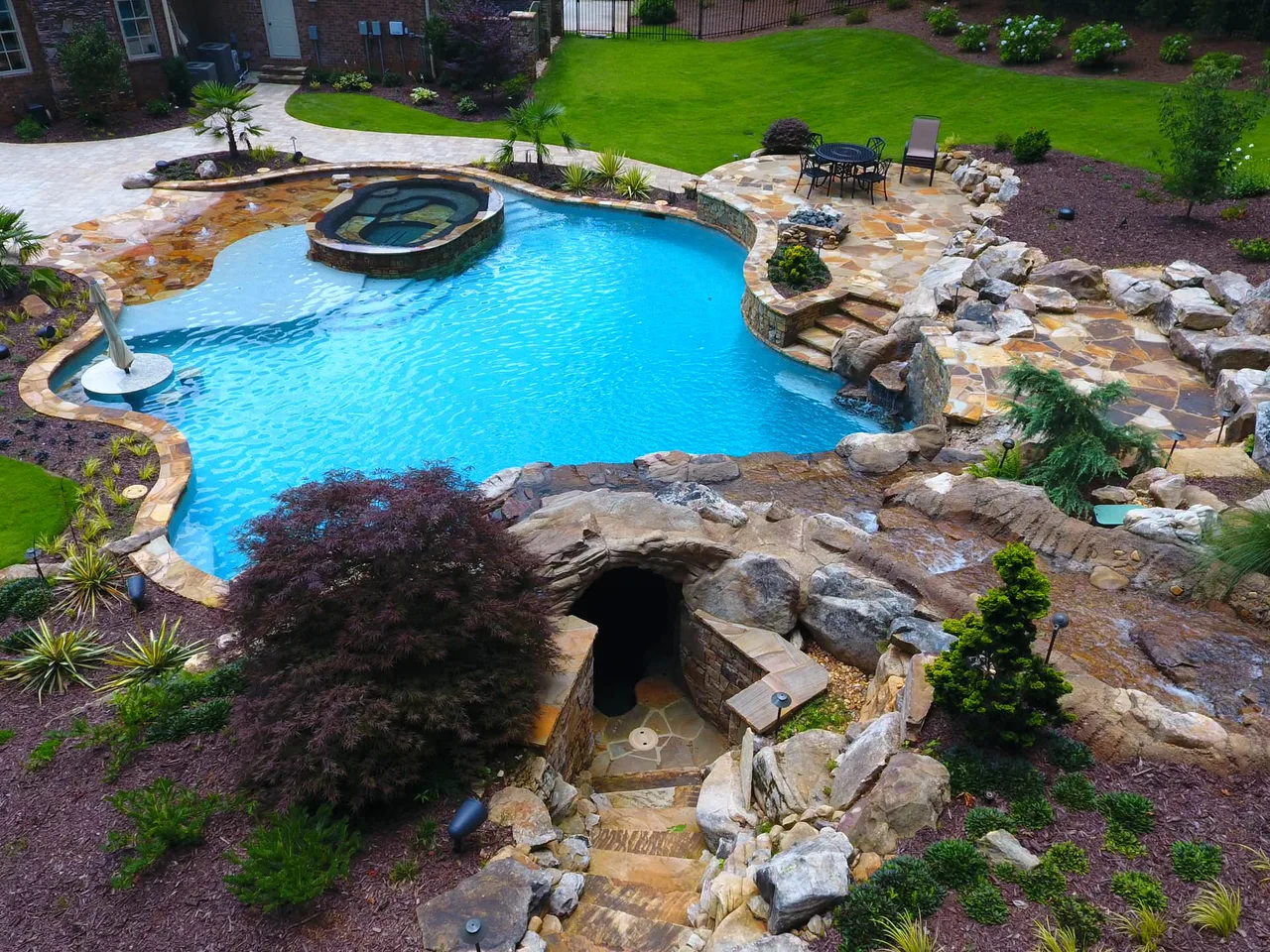 Aerial view of freeform pool with natural stone cave entrance, beach entry, spa, and layered rock waterfall feature surrounded by lush landscaping
