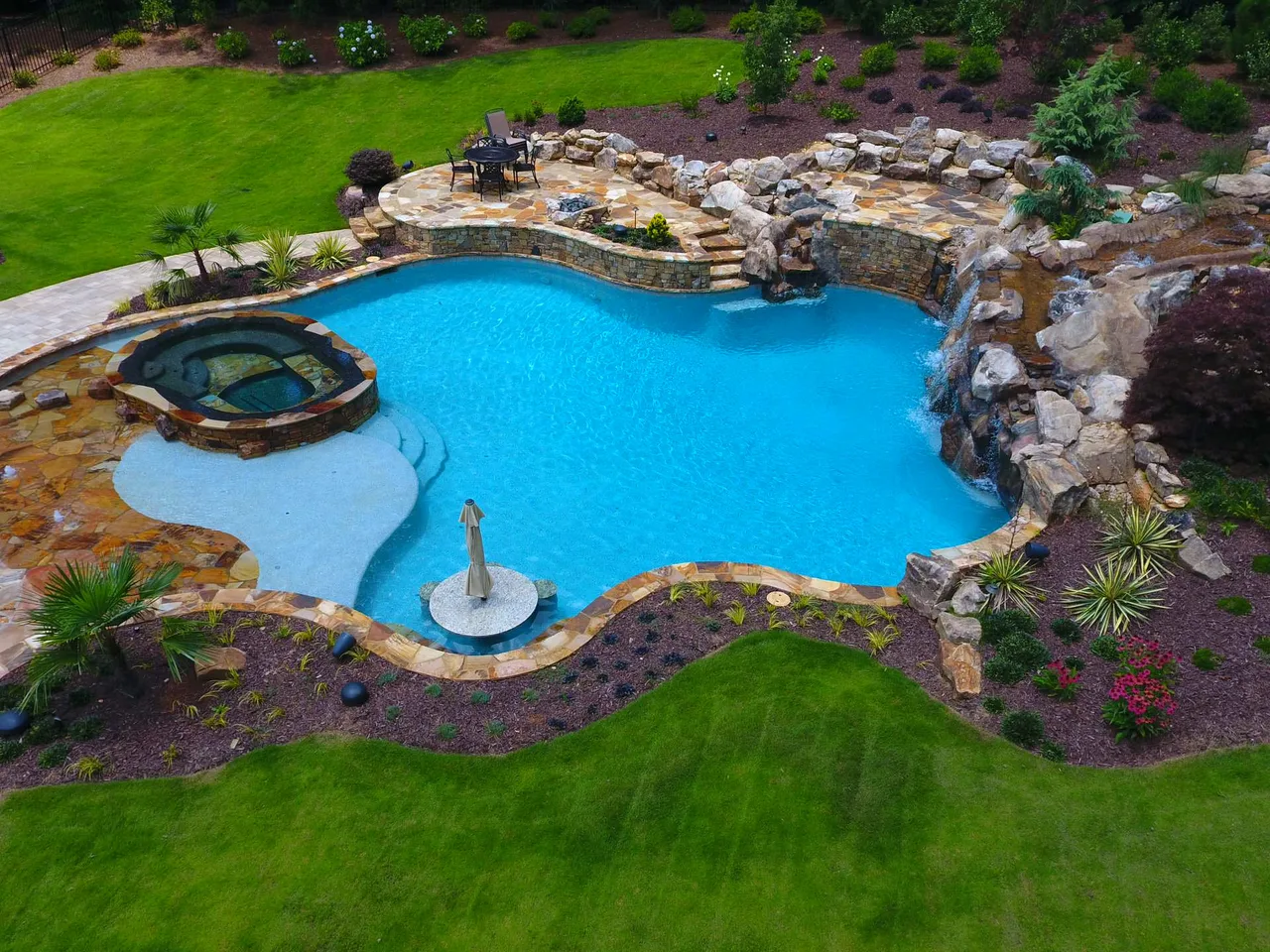 Aerial view of freeform pool with natural stone cave, beach entry, spa, and manicured lawn surrounding in Georgia