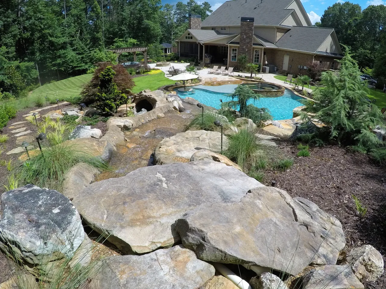 Aerial view of freeform pool with cave feature, beach entry, and natural boulders in manicured backyard