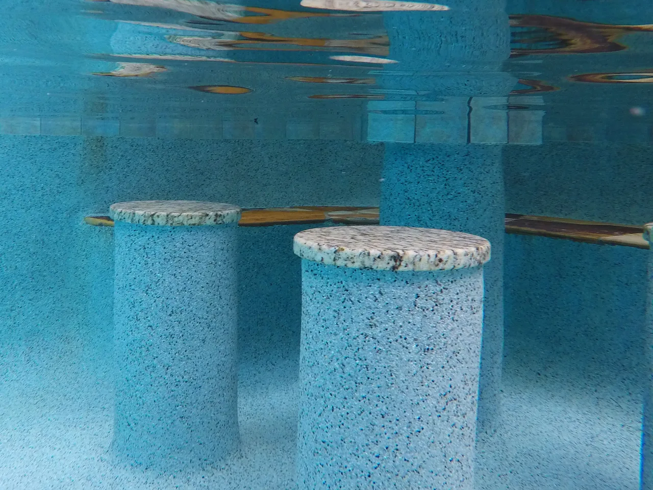 Underwater view of textured concrete pool seats and wooden handrails submerged in clear turquoise water