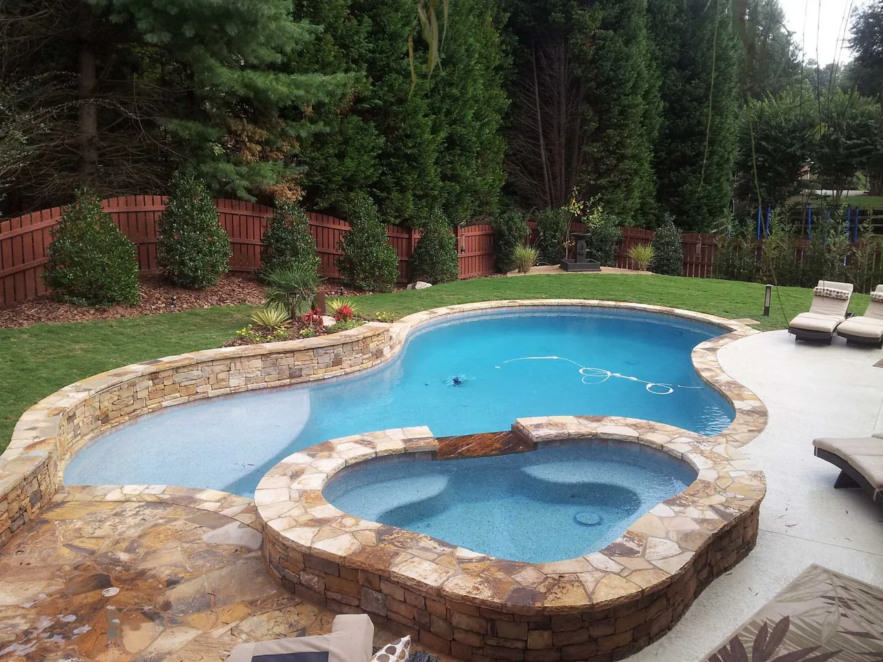 Freeform swimming pool with attached spa and stone coping, surrounded by manicured lawn and tall evergreen trees in Roswell, GA