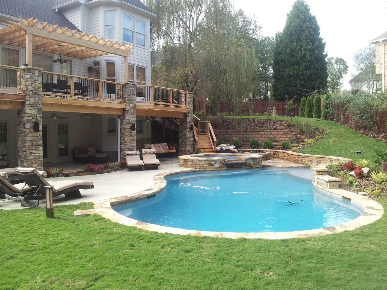 Curved custom pool with sun shelf and spa, stone coping, multi-level deck, and two-story home with pergola in Roswell, GA
