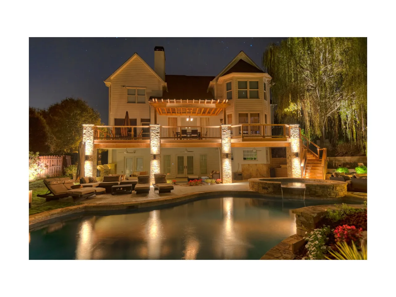 Illuminated custom pool with spa at dusk, stone pillars supporting wooden pergola, two-story home backdrop, Roswell, GA