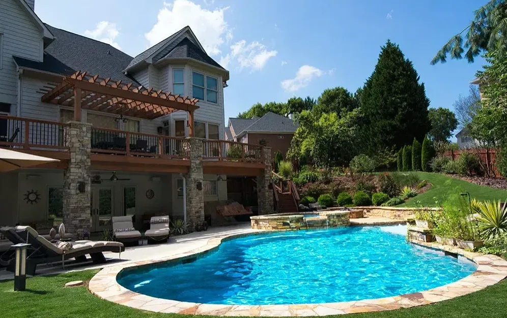 Curved freeform pool with blue water and light stone deck, spa to the left, multi-story stone and brick home in background under clear sky in Roswell, GA