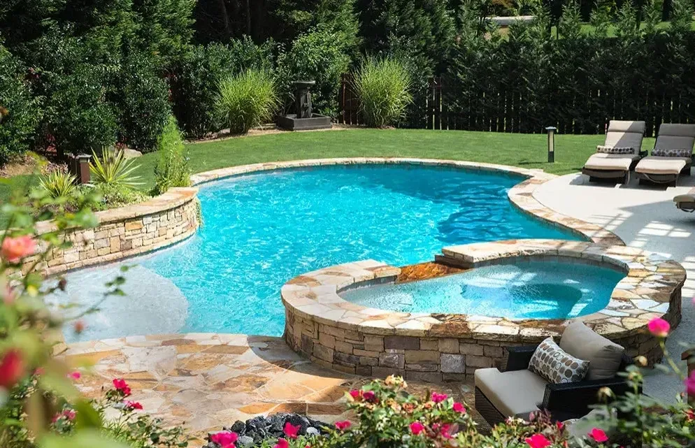 Freeform pool with attached spa, travertine deck, and natural stone spa surround overlooking manicured lawn and tree line in Roswell, GA