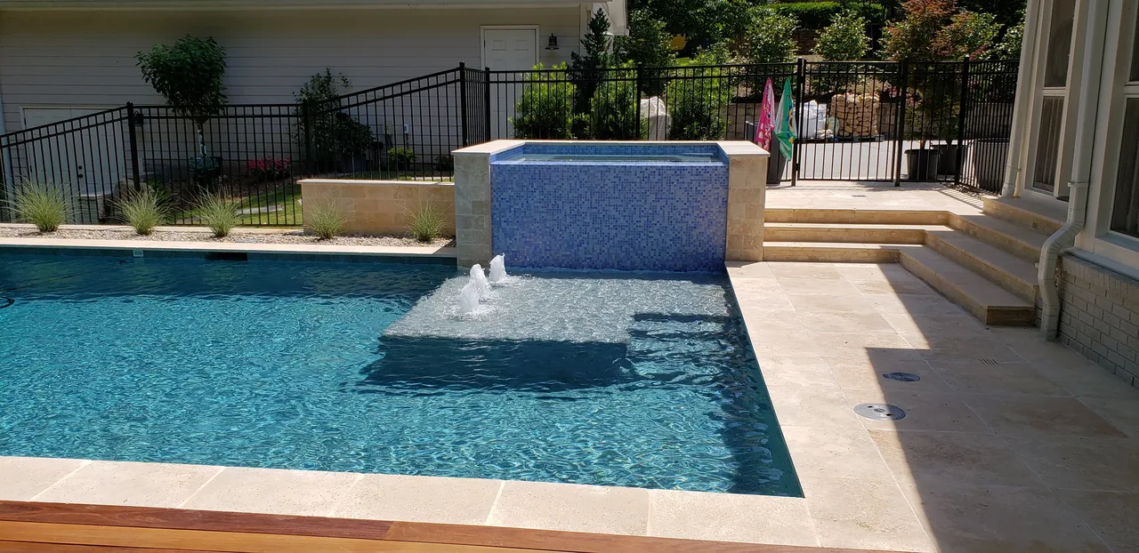 Modern linear pool with blue tile overflow spa and sheer descent water feature into clear turquoise water, travertine coping, and ipe wood deck