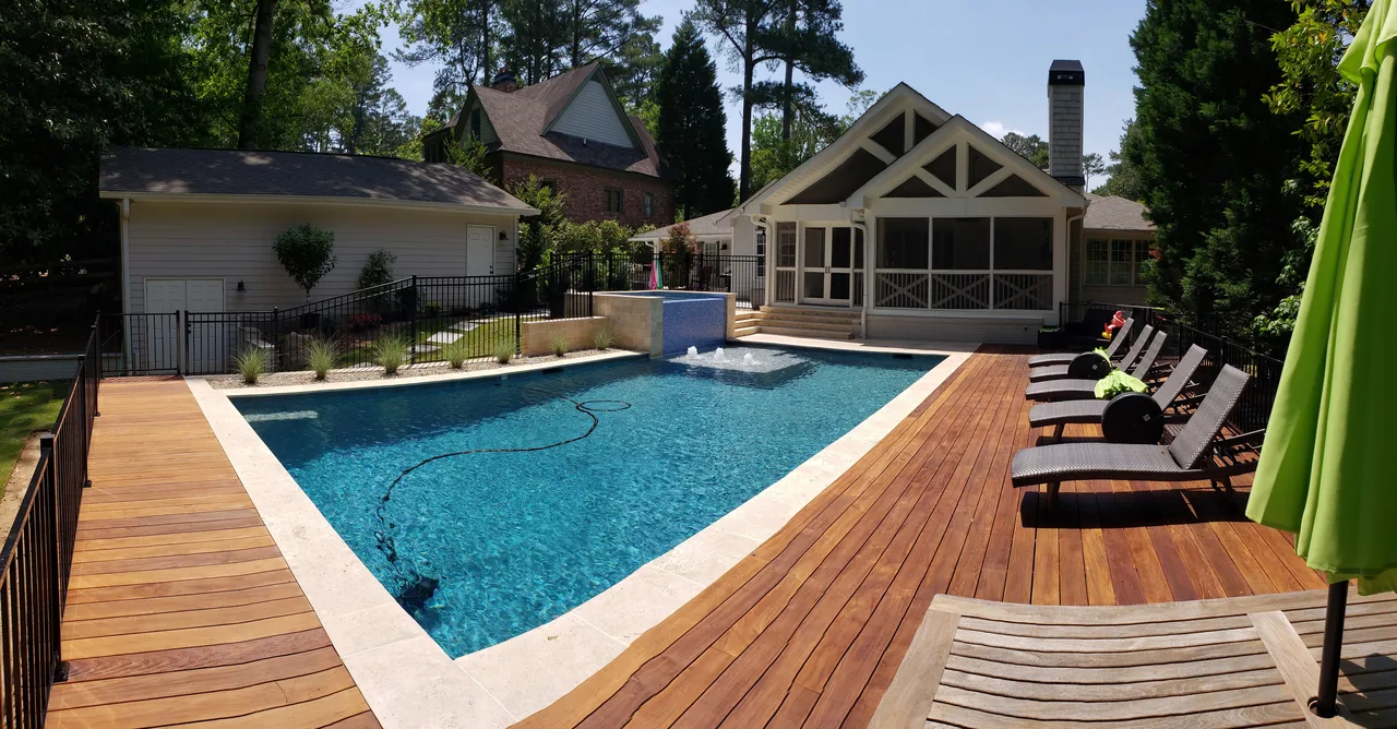 Modern linear pool with white travertine coping, clear blue water, ipe wood decking, lounge chairs, and screened pavilion in sunny Atlanta backyard