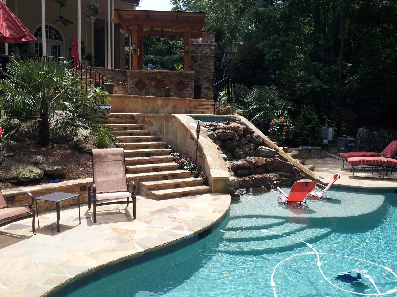 Curved pool deck with travertine pavers, natural stone water feature, and stepped entry leading to home in Suwanee, GA