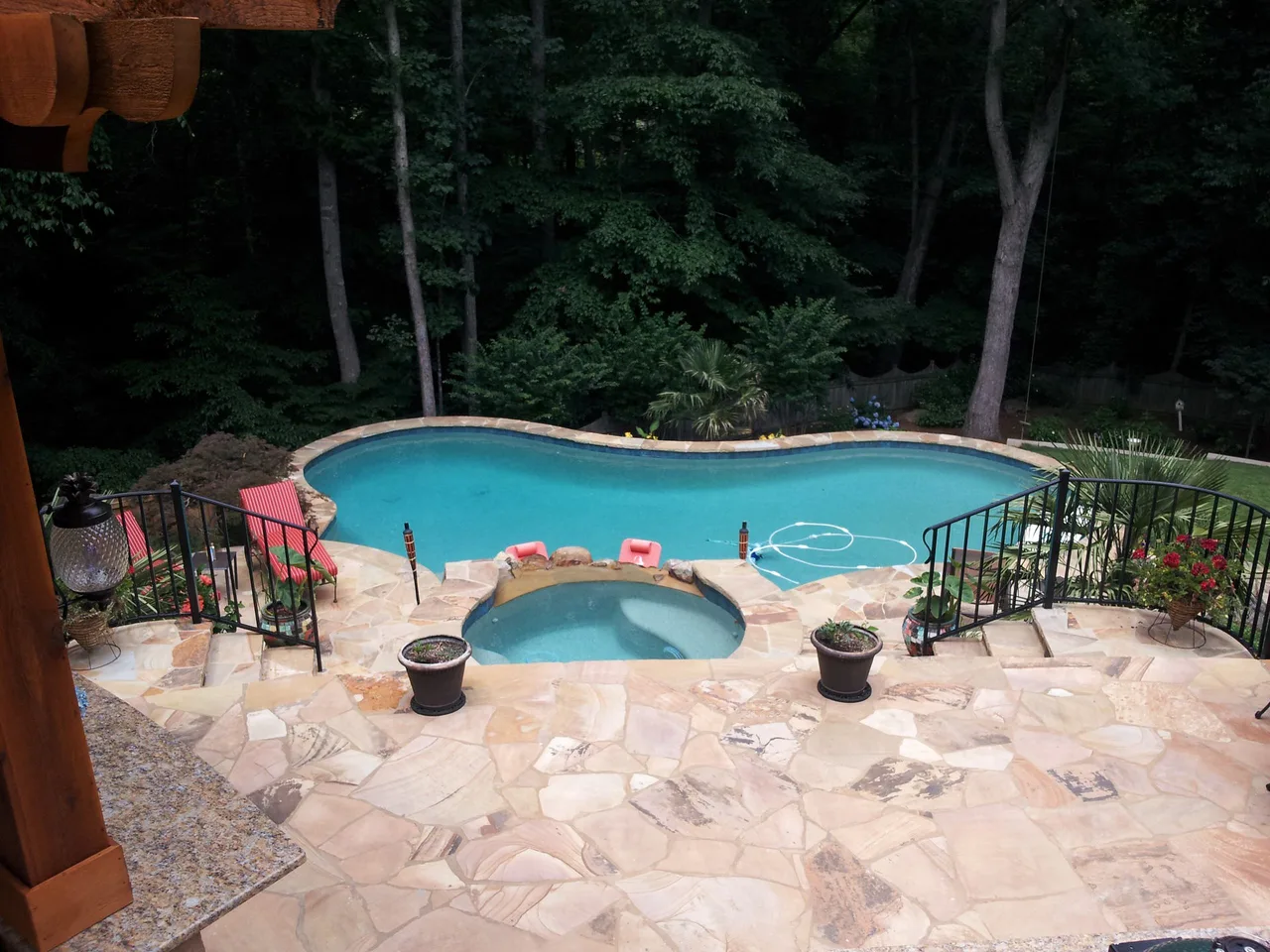 Elevated view of free-form pool with attached spa, travertine deck, black metal railings, and tall trees surrounding the backyard in Suwanee, GA