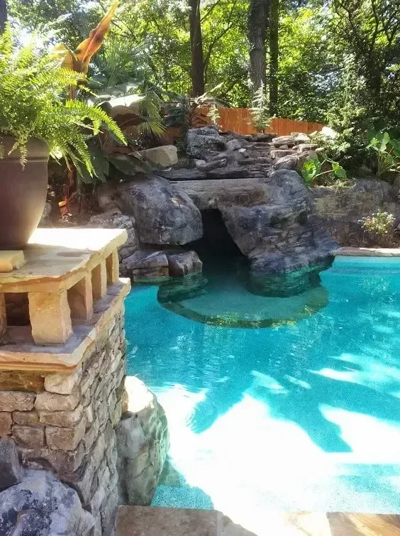 Tropical free-form pool with turquoise water, natural rock waterfall, stone deck, and dense green vegetation overhead