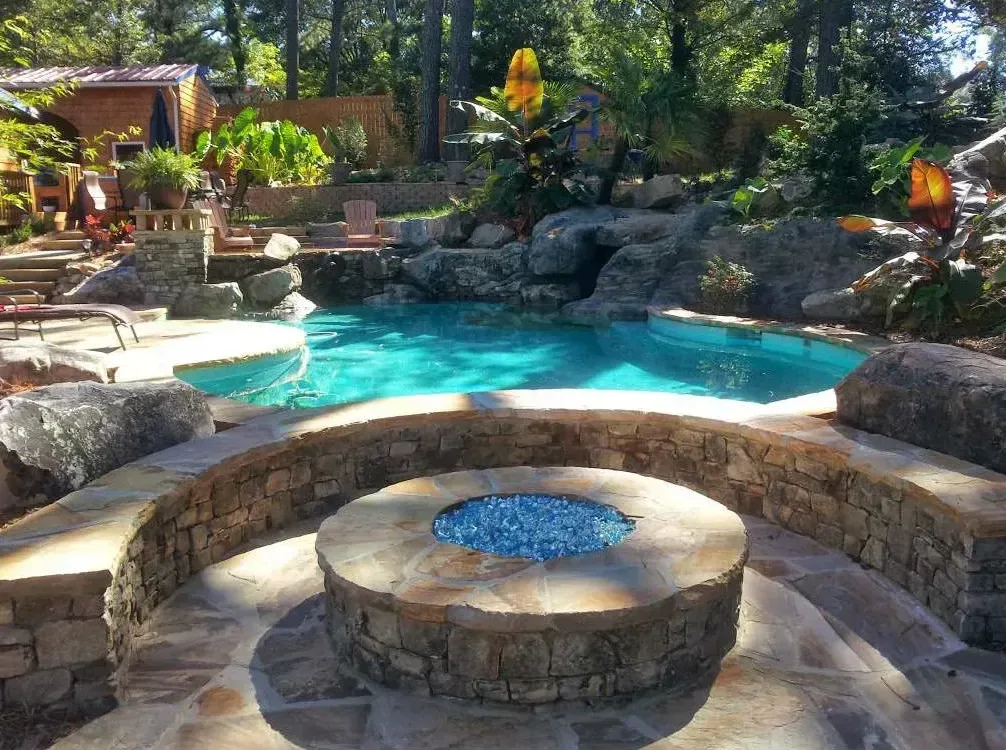 Circular stone fire bowl on travertine deck overlooking turquoise free-form pool with natural rock landscaping
