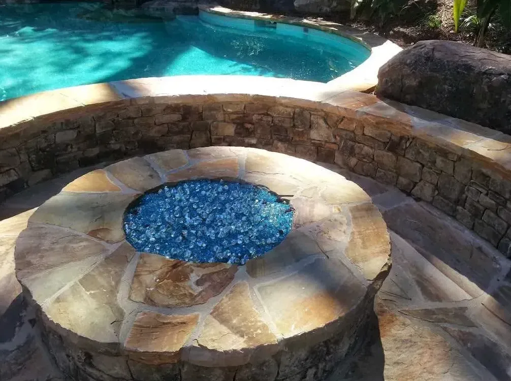 Stone fire bowl with blue glass gems on travertine deck beside free-form pool with turquoise water