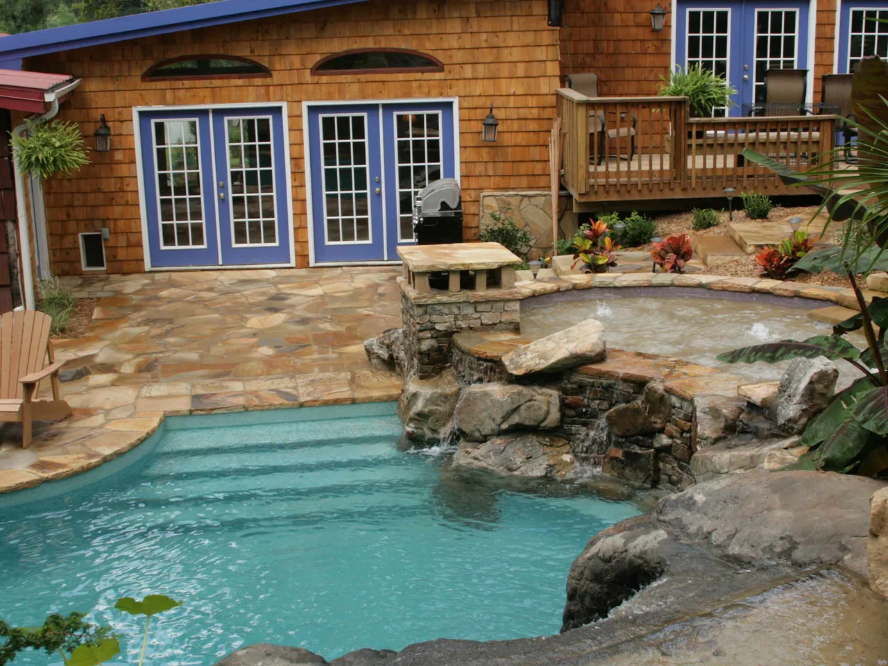 Free-form pool with natural stone waterfall, turquoise water, and travertine deck adjoining cedar-sided home with blue doors
