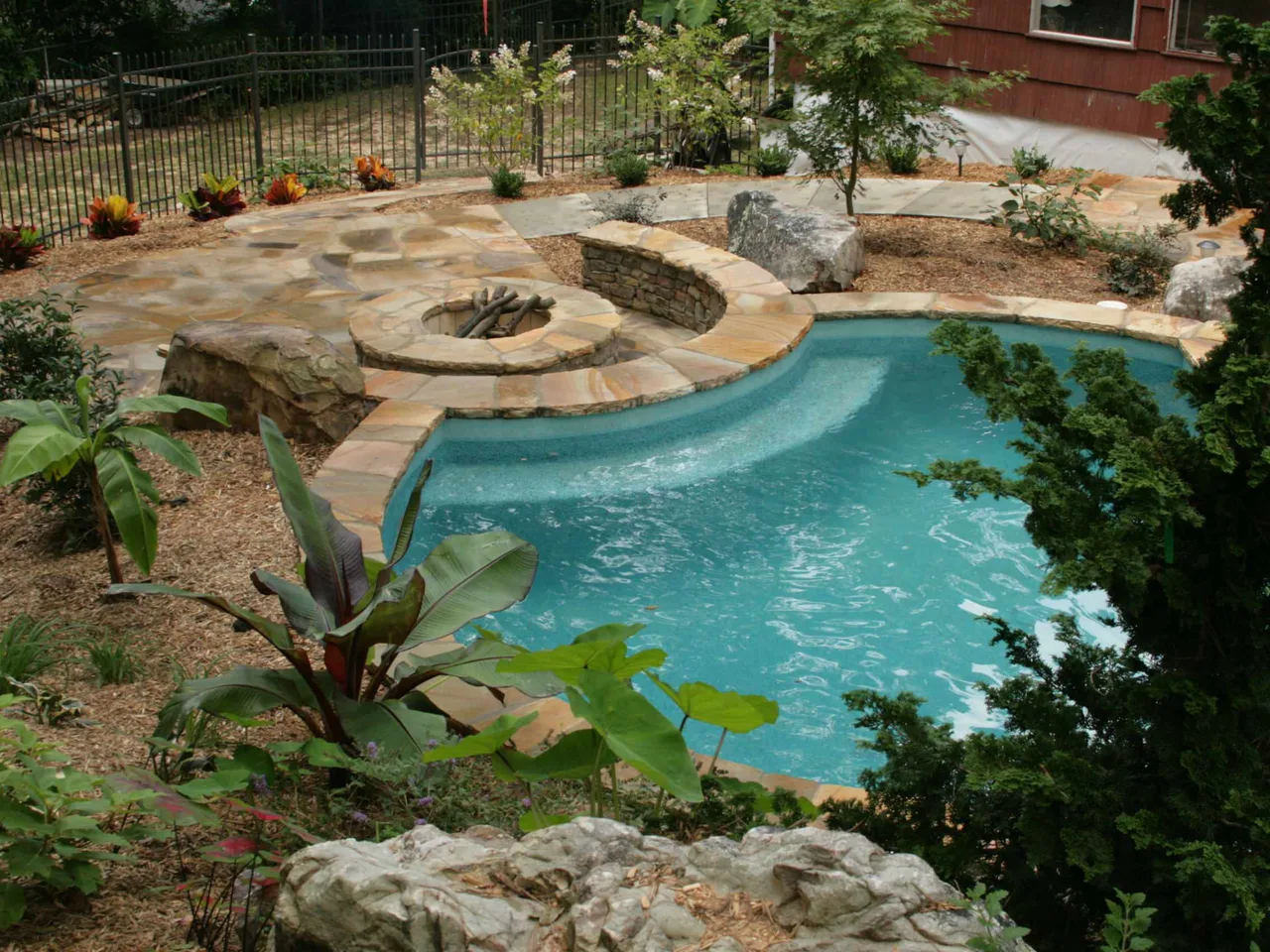 Free form pool with curved travertine deck, fire bowl feature, and natural stone boulders surrounded by tropical landscaping