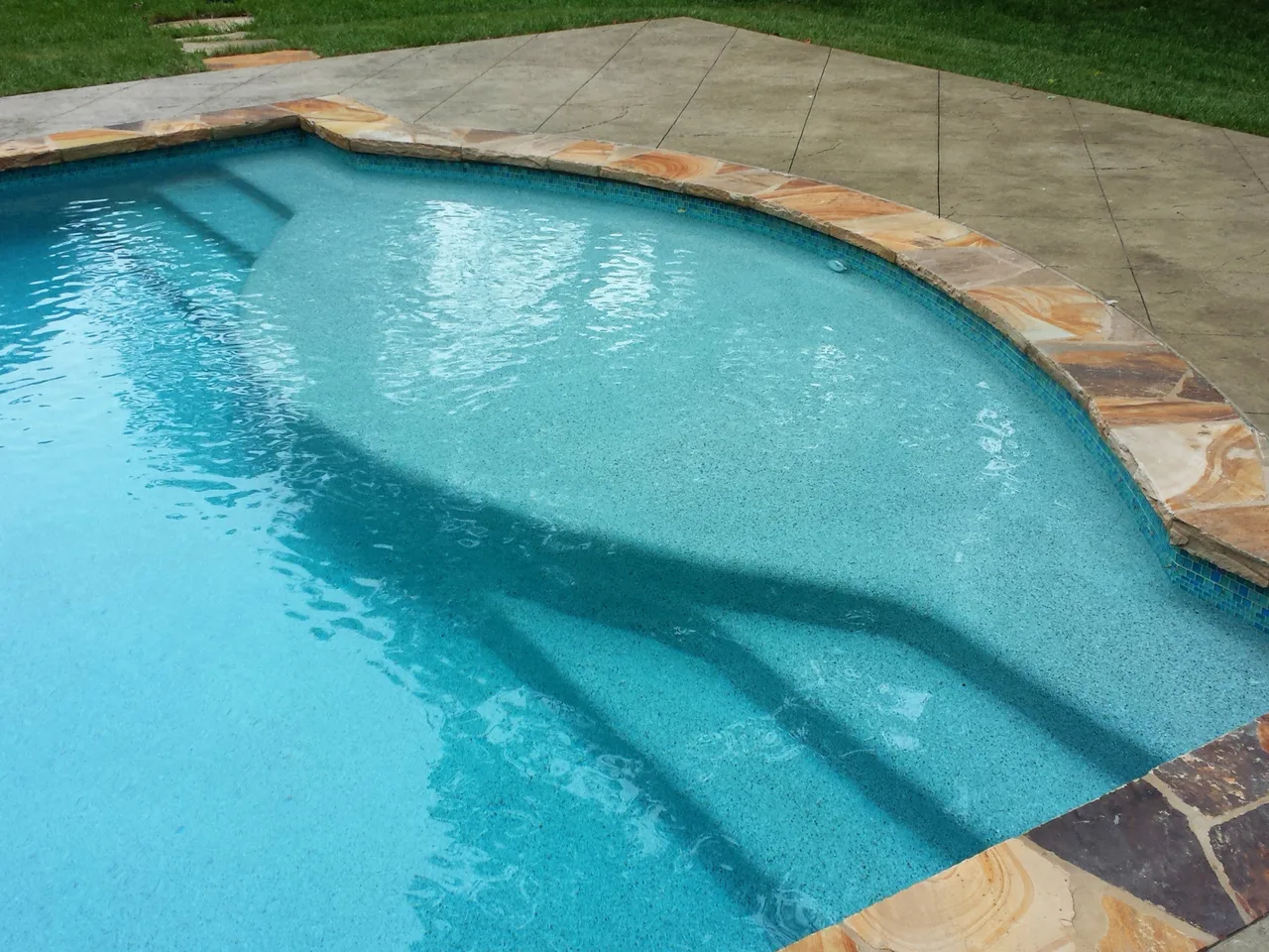 Curved custom pool with turquoise water and sun shelf, natural stone border, and manicured lawn in Atlanta, GA