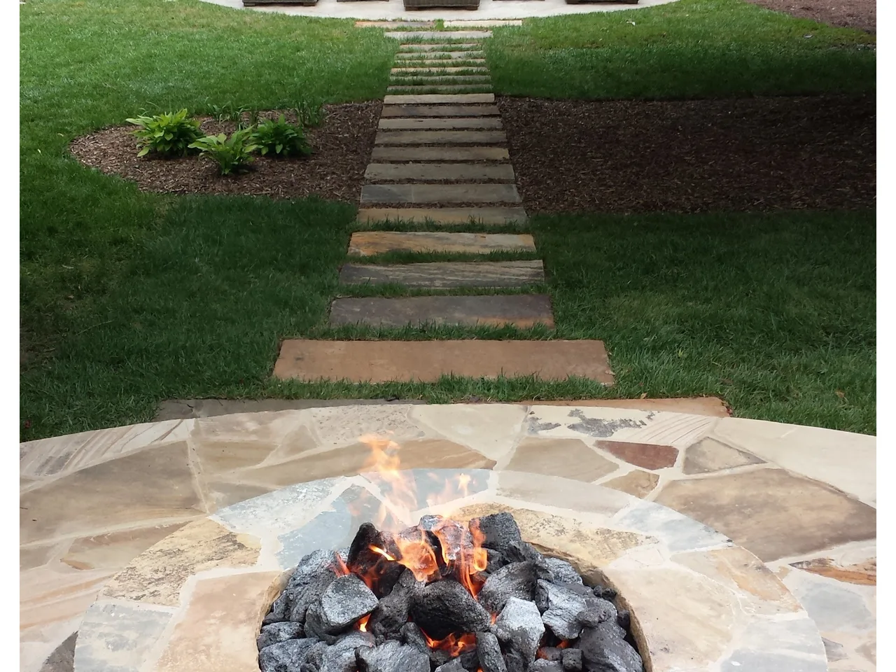 Stone fire pit with active flames on travertine deck, straight pathway and manicured lawn leading to pool in Atlanta, GA