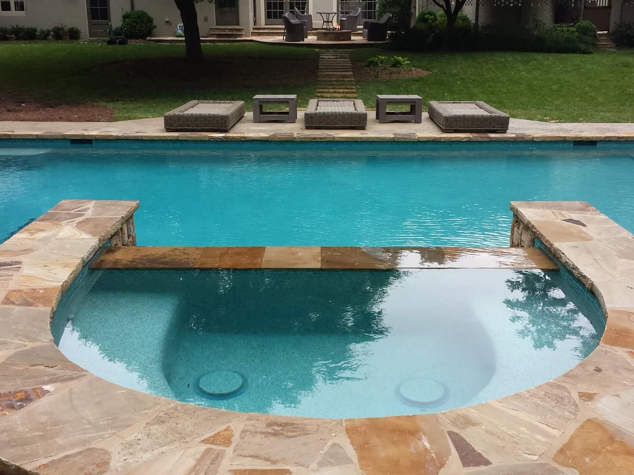 Curved spa with stone decking overlooking large rectangular pool with travertine deck and manicured lawn in Atlanta, GA