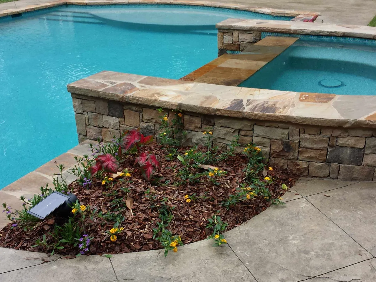 Turquoise pool with stone sun shelf, spa, and stone retaining wall landscaped with colorful flowering plants in Atlanta, GA